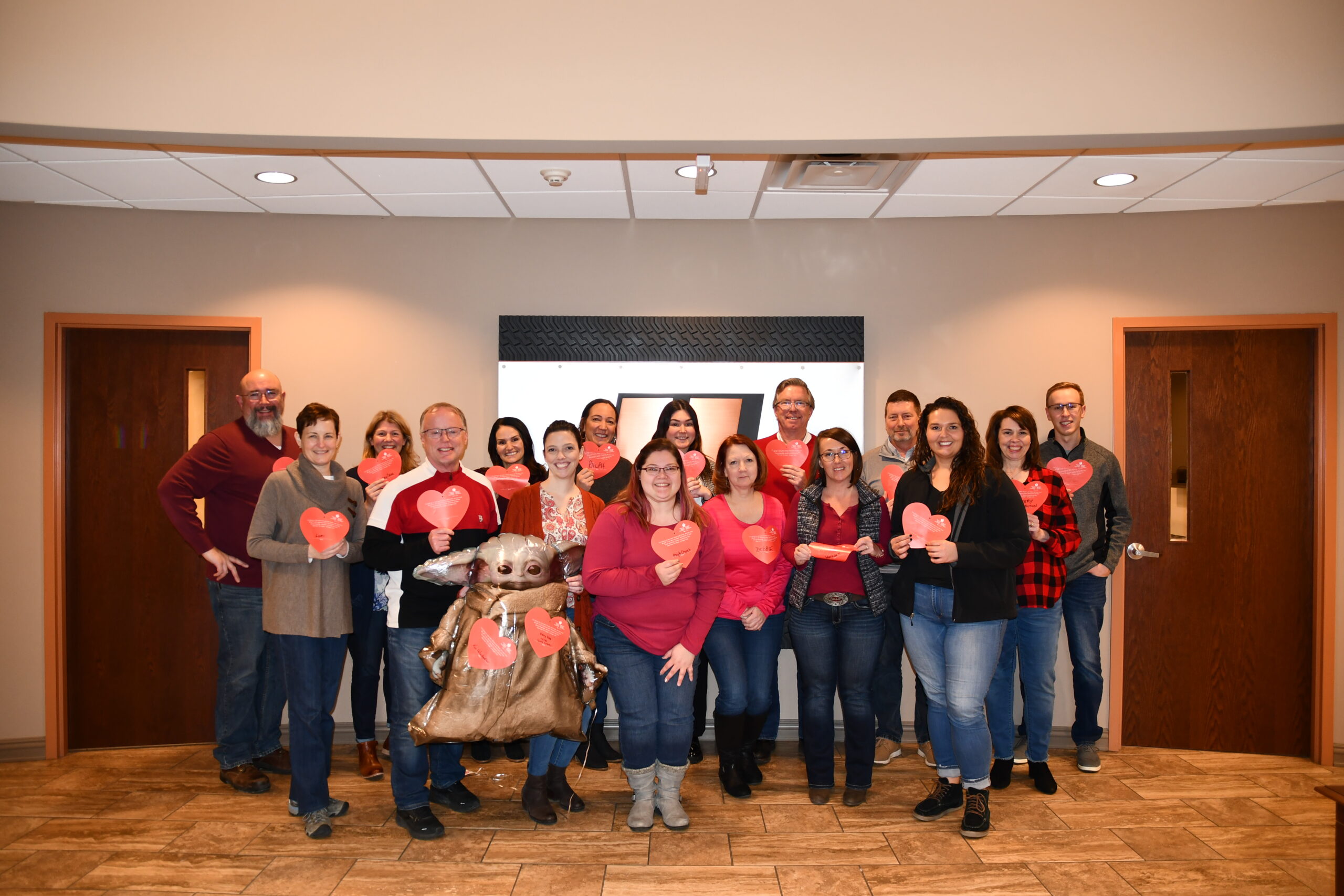 A group of Hunter Truck employees gather in the front office, they are smiling and wearing red clothing. They hold paper cut out hearts.